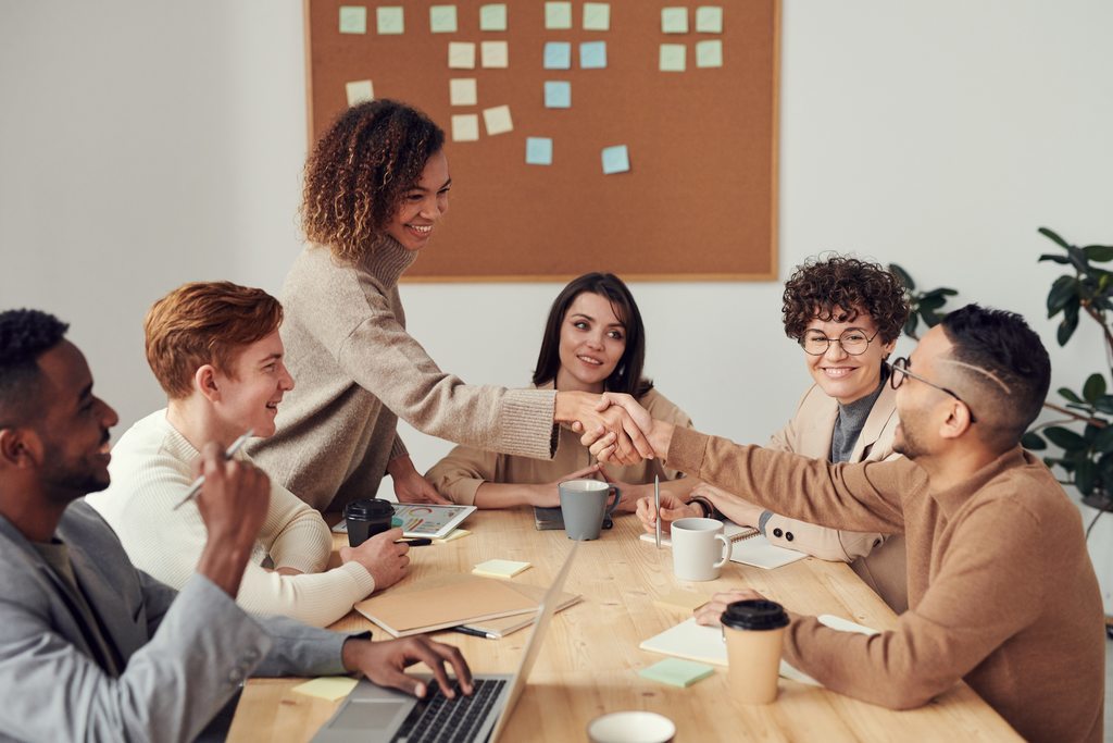Quadro com post its ao fundo, em primeiro plano grupo de pessoas reunidas à mesa de reunião. Um homem e uma mulher estão fazendo um cumprimento, apertando as mãos. Representando um acordo sobre benefícios flexíveis