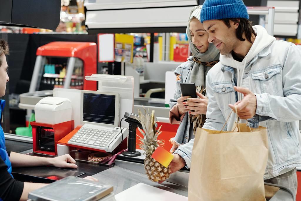 Homem segurando sacola e abacaxi, e mulher segurando celular. O casal está passando compras em caixa de supermercado, utilizando desconto com vale alimentação