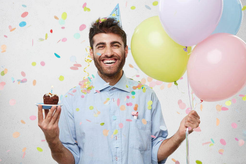 Funcionário feliz, com chapéu de festa, segurando balões em uma mão e um pequeno bolo na outra.
