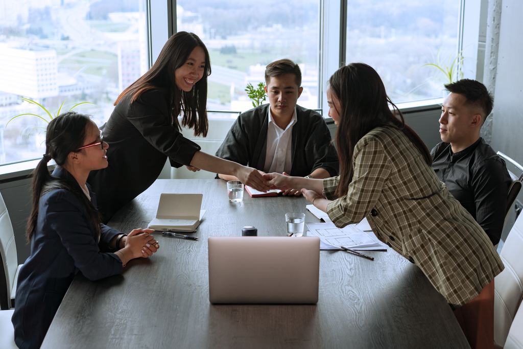 Grupo de cinco pessoas de roupa social em reunião de trabalho, ao redor de uma mesa que abriga um notebook, duas agendas, documentos e copos de água. Duas pessoas estão em pé, sorrindo se cumprimentando, simbolizando satisfação no trabalho