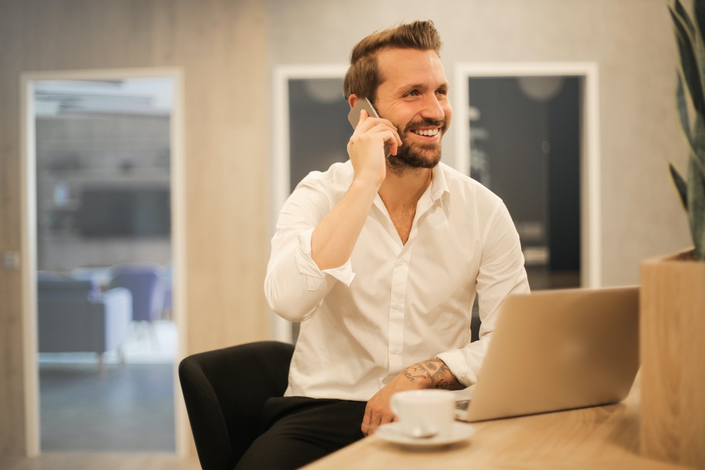 Simbolizando tipos de ajuda de custa: Um colaborador vestido com camisa branca social sorrindo falando ao celular, e um notebook a sua frente sobre a mesa e uma xícara ao lado do notebook